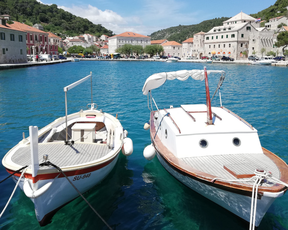 Two traditional wooden boats floating in a clear blue harbor, with a scenic coastal town featuring stone buildings and red rooftops in the background.