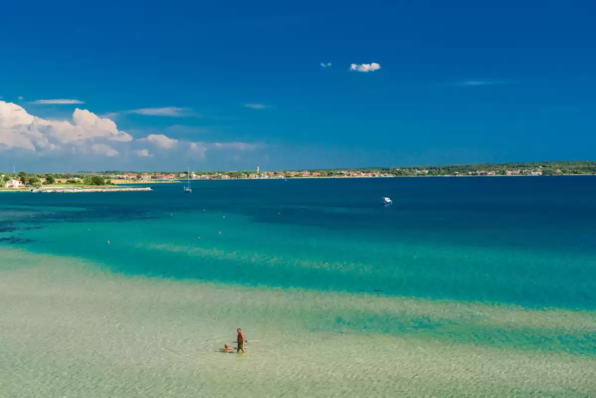 Blick auf den Sandstrand Sabunike mit türkisfarbenem seichtem Meer und Küste im Hintergrund unter klarem blauen Himmel, ideal für einen Familienurlaub.