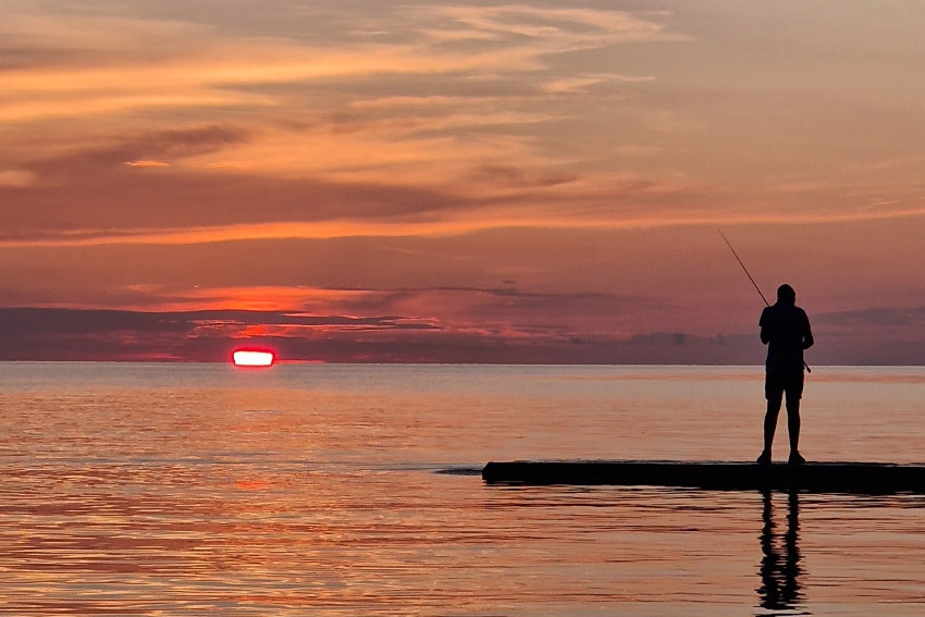 Silhouette eines Fischers auf einem Pier bei Sonnenuntergang mit warmen orange-roten Reflexionen auf dem ruhigen Meer.