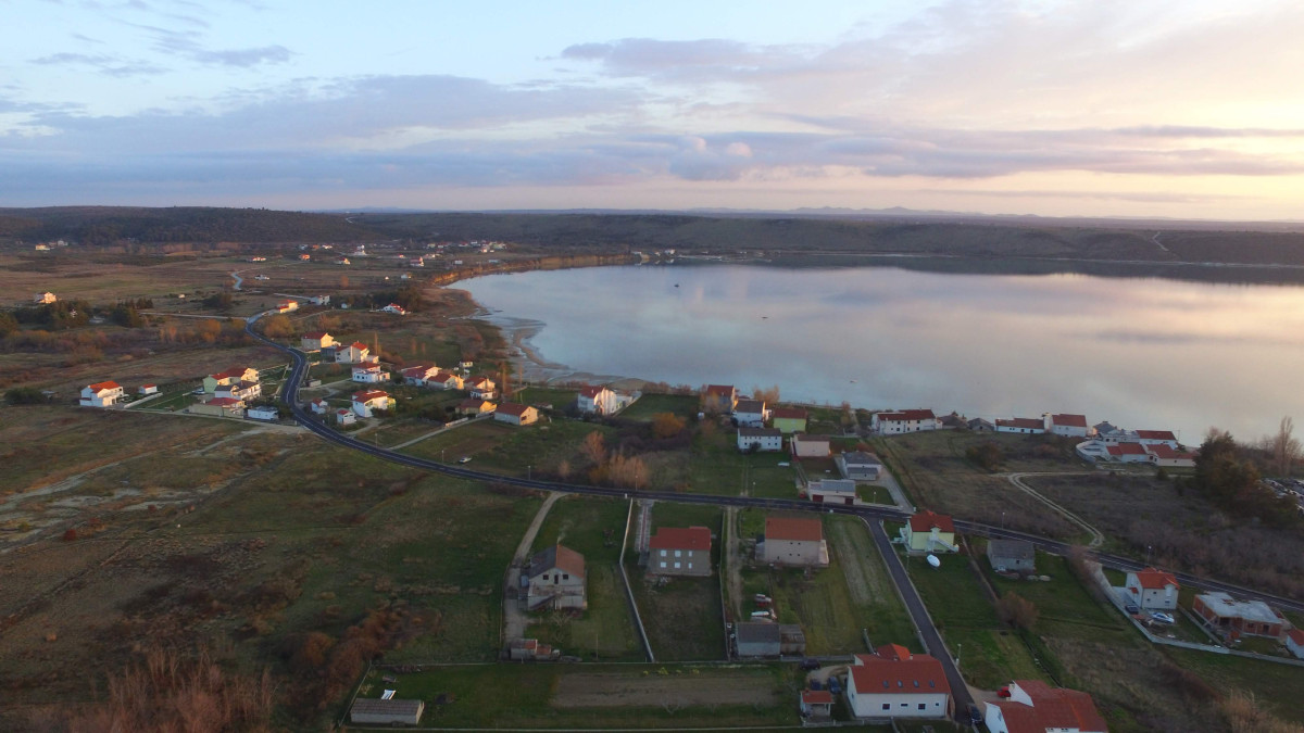 Luftaufnahme des Ljubač-Strandes, einer ruhigen und flachen Bucht, umgeben von Häusern, grünen Feldern und einer malerischen Landschaft unter einem sanften Sonnenuntergangshimmel.